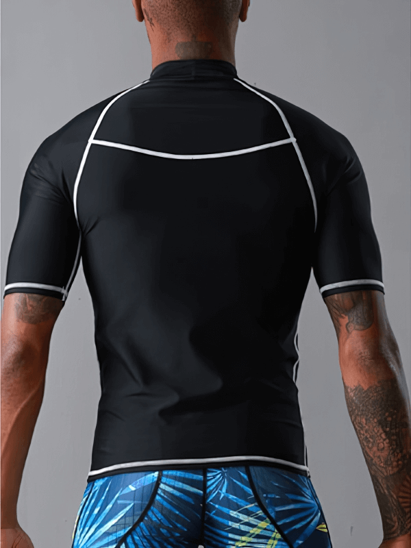 Man wearing a short sleeve sports elastic beach T-shirt for water sports, in black with white piping, and patterned blue swim shorts, back view.